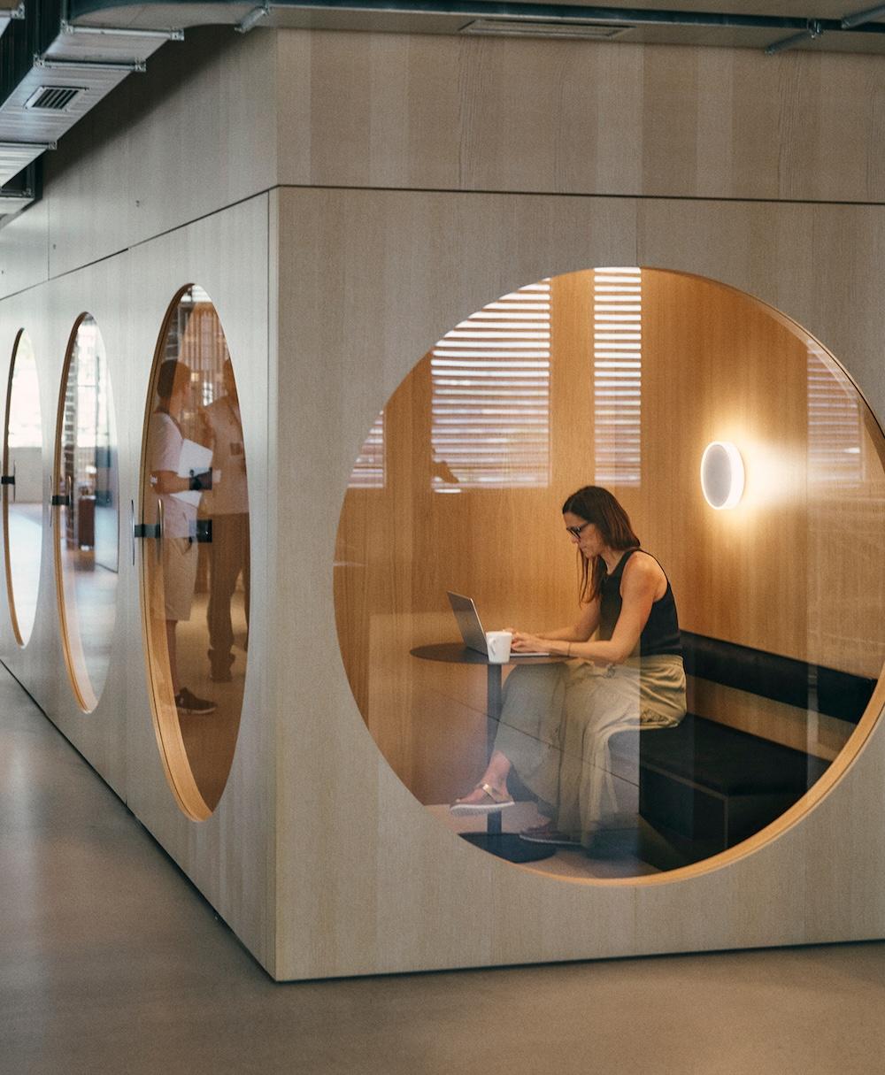 A woman sits at a small table inside a circular-windowed wooden office pod, working on a laptop with a mug nearby and a wall sconce behind her.