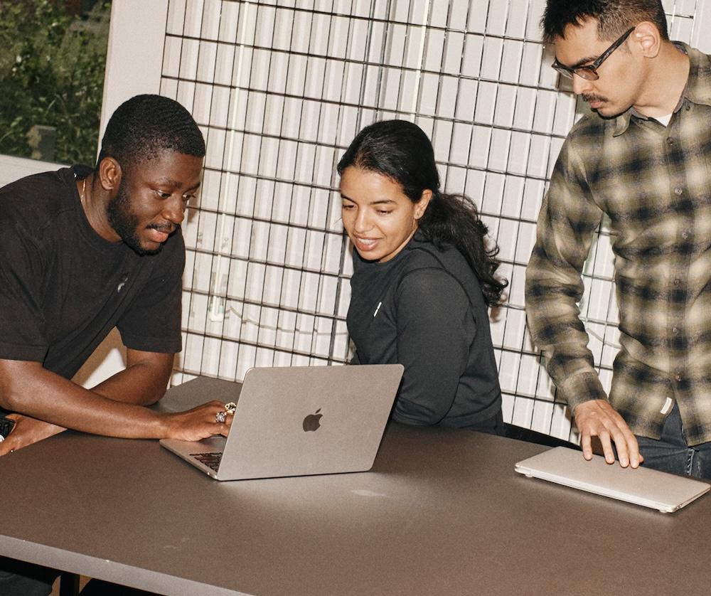 Three people lean over a table and look at an open laptop while a closed laptop sits nearby.
