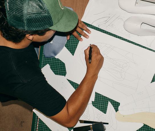 Person wearing a green cap sketches shoe pattern templates on paper spread over a green cutting mat with shoe components nearby.