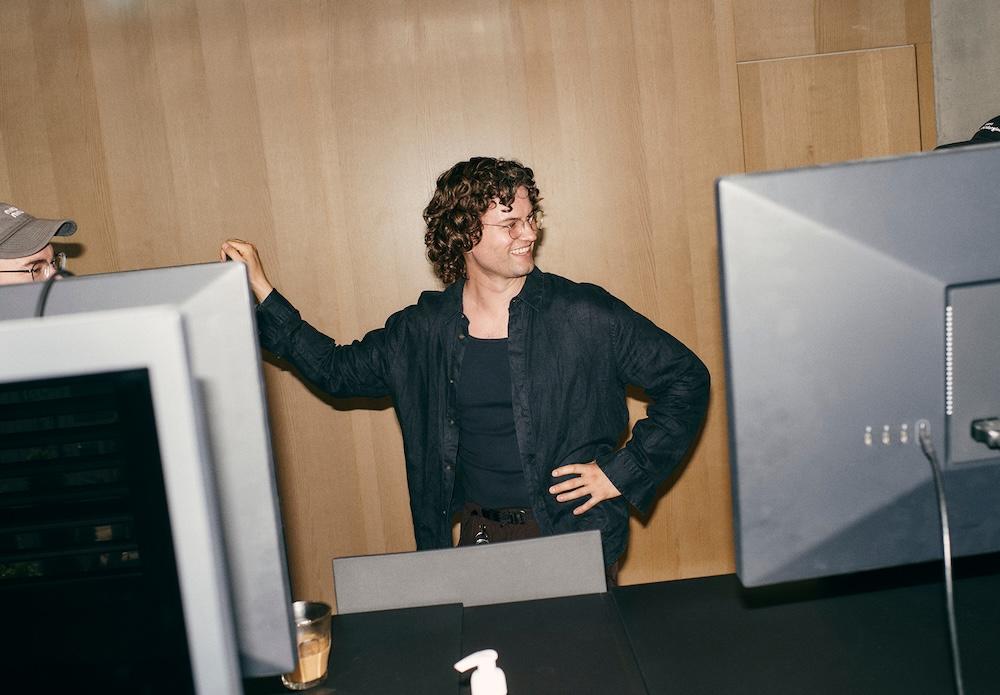 A smiling man with curly hair and glasses stands with one hand on his hip between computer monitors against a wood-paneled wall.