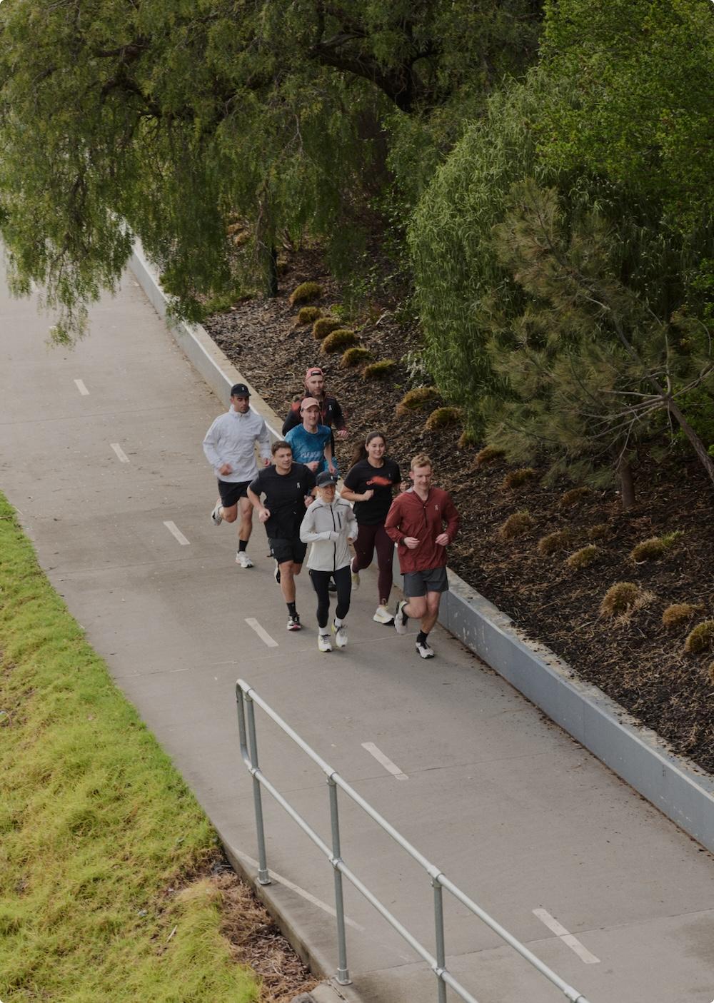 A group of eight people jogging together on a paved path beside a planted embankment and trees.