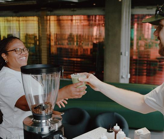 A smiling person behind a counter hands a small cup of green drink to a customer, with a coffee grinder and condiment bottles visible.