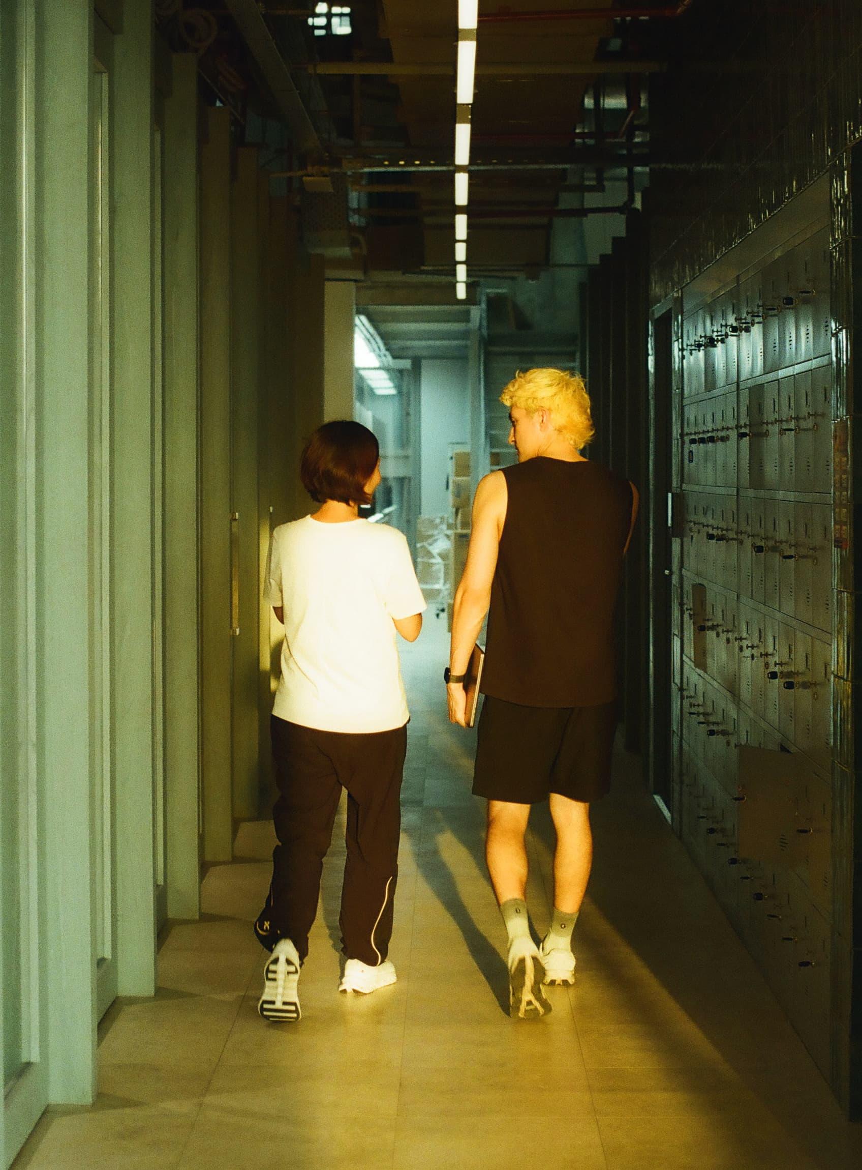 Two people walk away from the camera down a narrow, fluorescent-lit hallway lined with lockers.