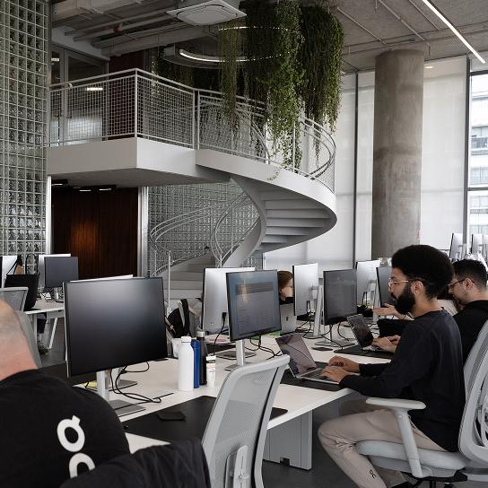 Open-plan office where people work at rows of desks with desktop monitors and laptops, with a white spiral staircase and hanging greenery in the background.