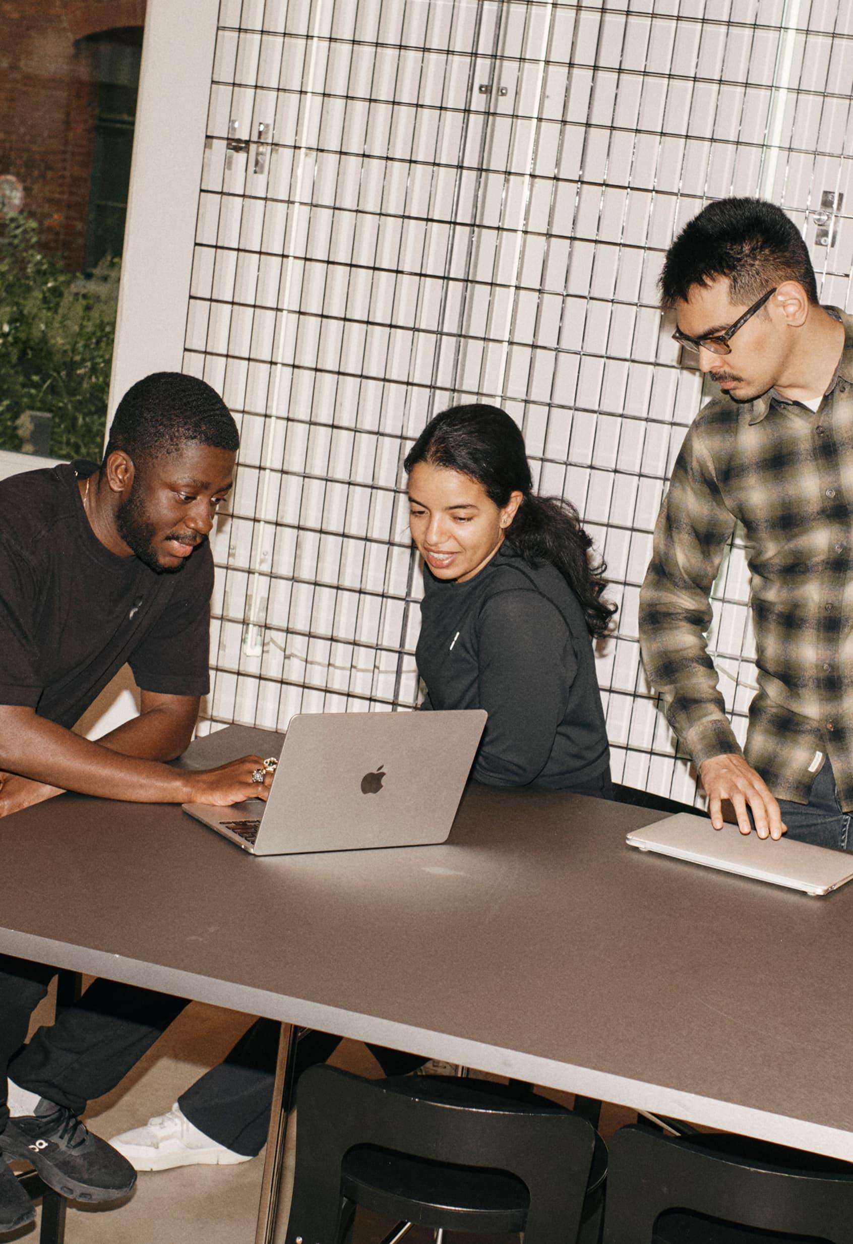 Three colleagues gather around a laptop, reviewing work together in a well-lit meeting space.