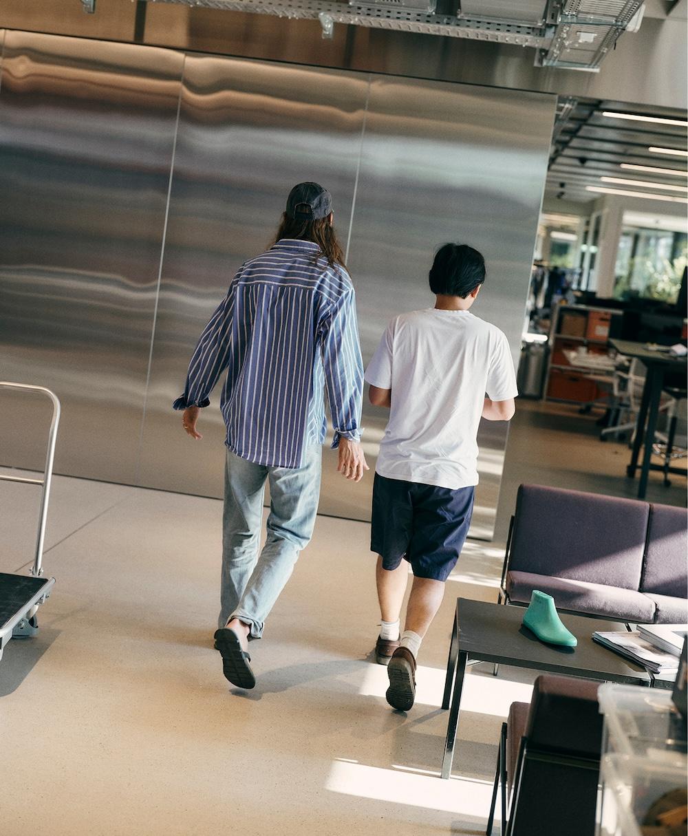 Two people walk away from the camera through an office interior, one wearing a striped long-sleeve shirt, cap and jeans and the other wearing a white T-shirt and dark shorts.