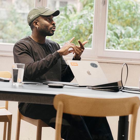 A man wearing a green baseball cap and dark long-sleeve shirt sits at a table with an open laptop and a glass of water, gesturing with his hands in front of a window.