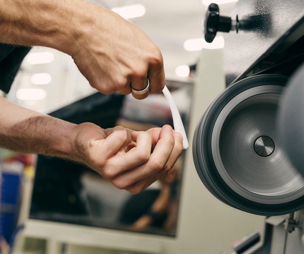 Two hands holding a thin white strip of material against a spinning grinding wheel on a machine.