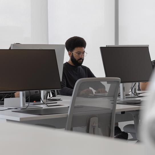 A person wearing glasses sits at a desk in an office, typing on a keyboard surrounded by multiple large computer monitors.