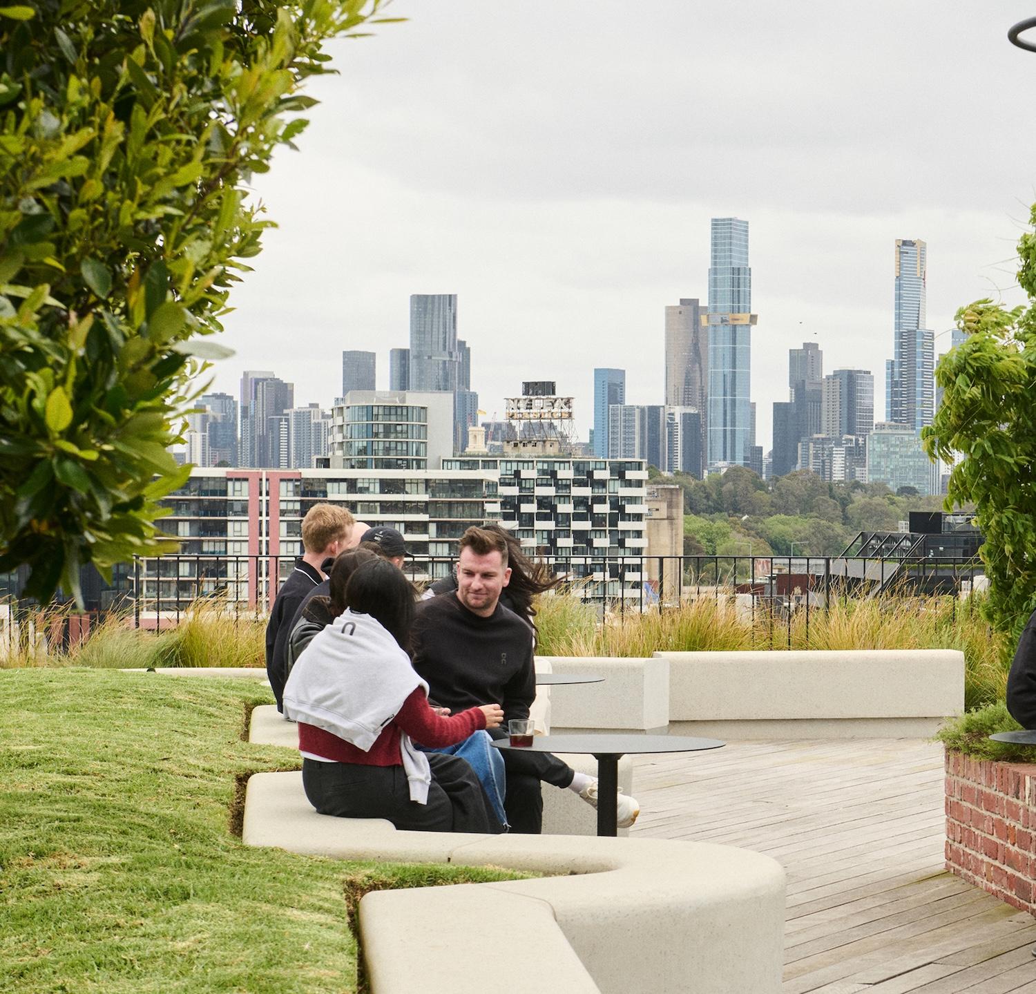 Group of people sit and talk at curved concrete benches on a rooftop garden with a city skyline in the background.