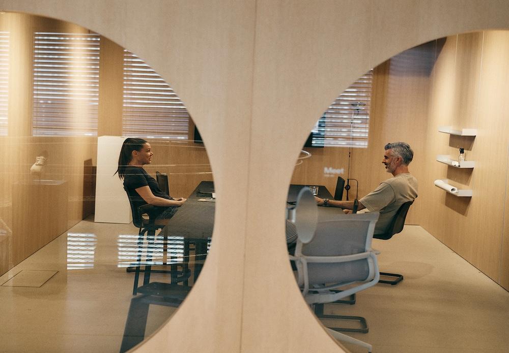 Two people sit across from each other at a table in a wood-paneled meeting room, seen through a circular opening in a partition.