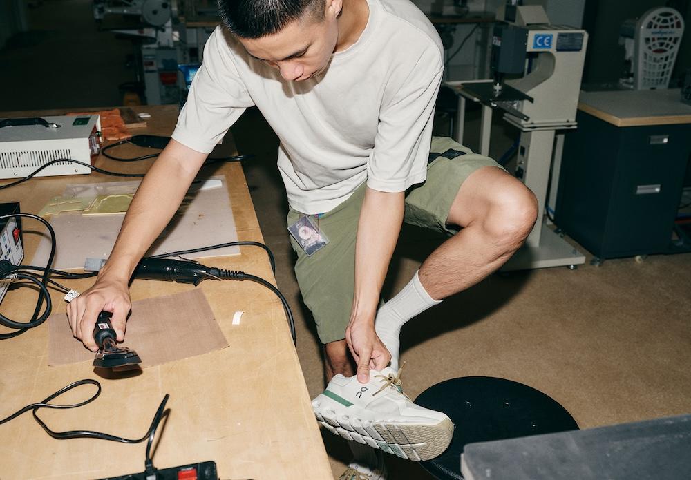 A person in a white T-shirt and green shorts leans over a workbench using a handheld power tool on a piece of material while holding a white sneaker.