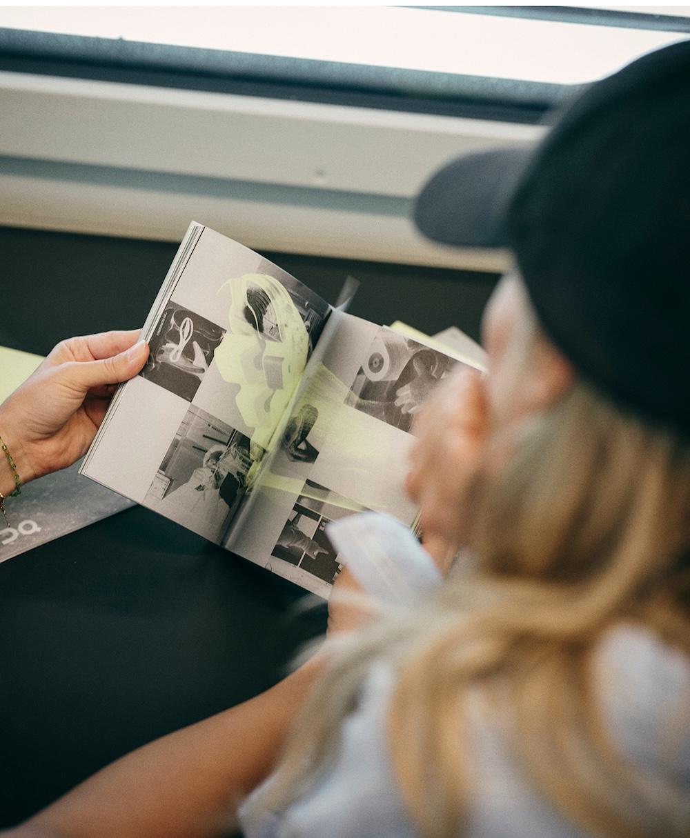 A person wearing a dark cap looks at an open booklet of black-and-white photos with a translucent yellow overlay held by another person's hands near a window.