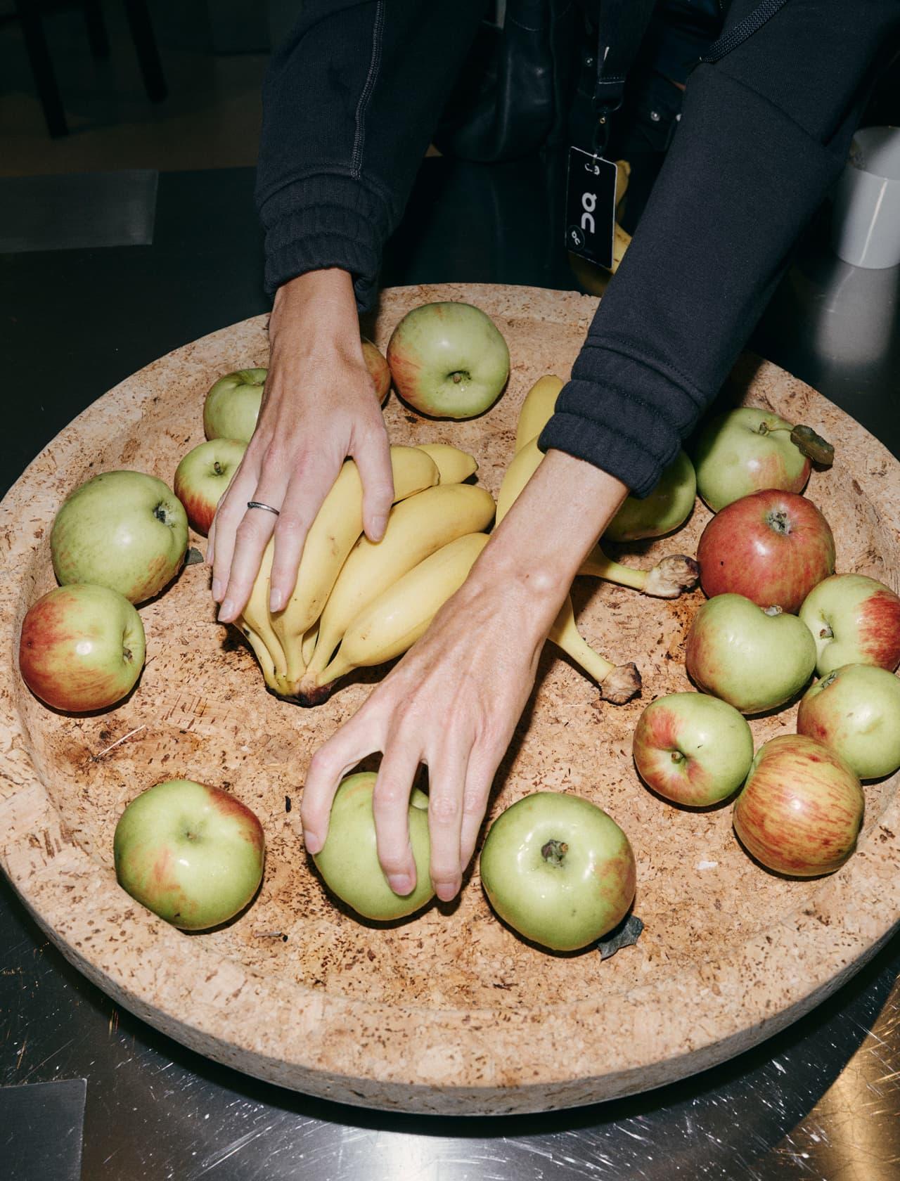 Two hands reach into a large shallow wooden bowl filled with bananas and green and red apples.