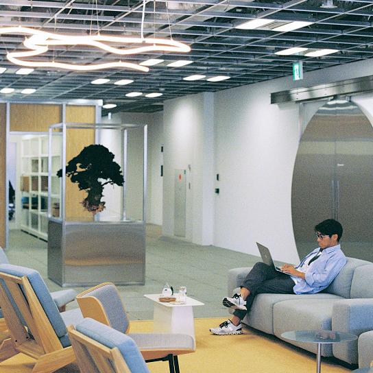 A person sits on a gray sofa using a laptop in a modern open office lounge with low seating, a glass-encased plant display, and curving ceiling lights.