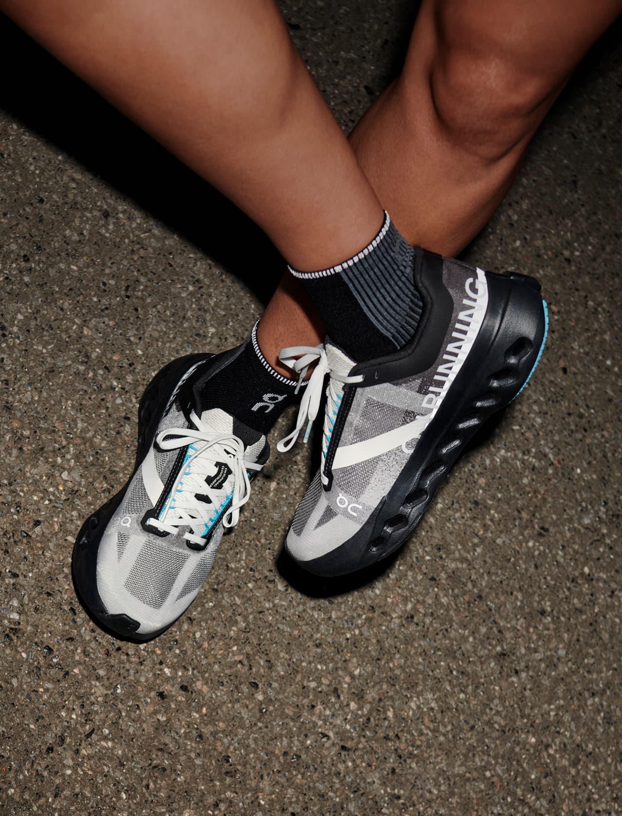 Crossed legs wearing gray and black running shoes with white laces and black ankle socks on textured concrete pavement.