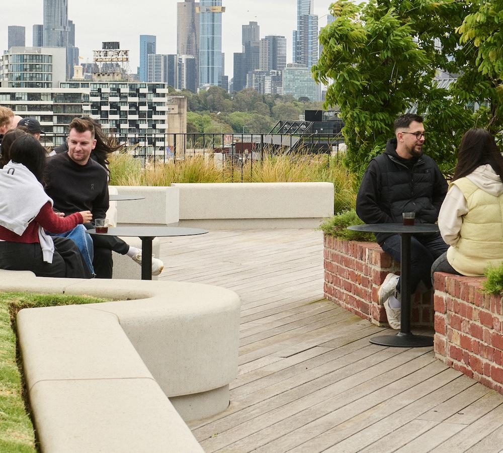 Several people sit and converse at small round tables on a rooftop terrace with planted beds and a city skyline in the background.