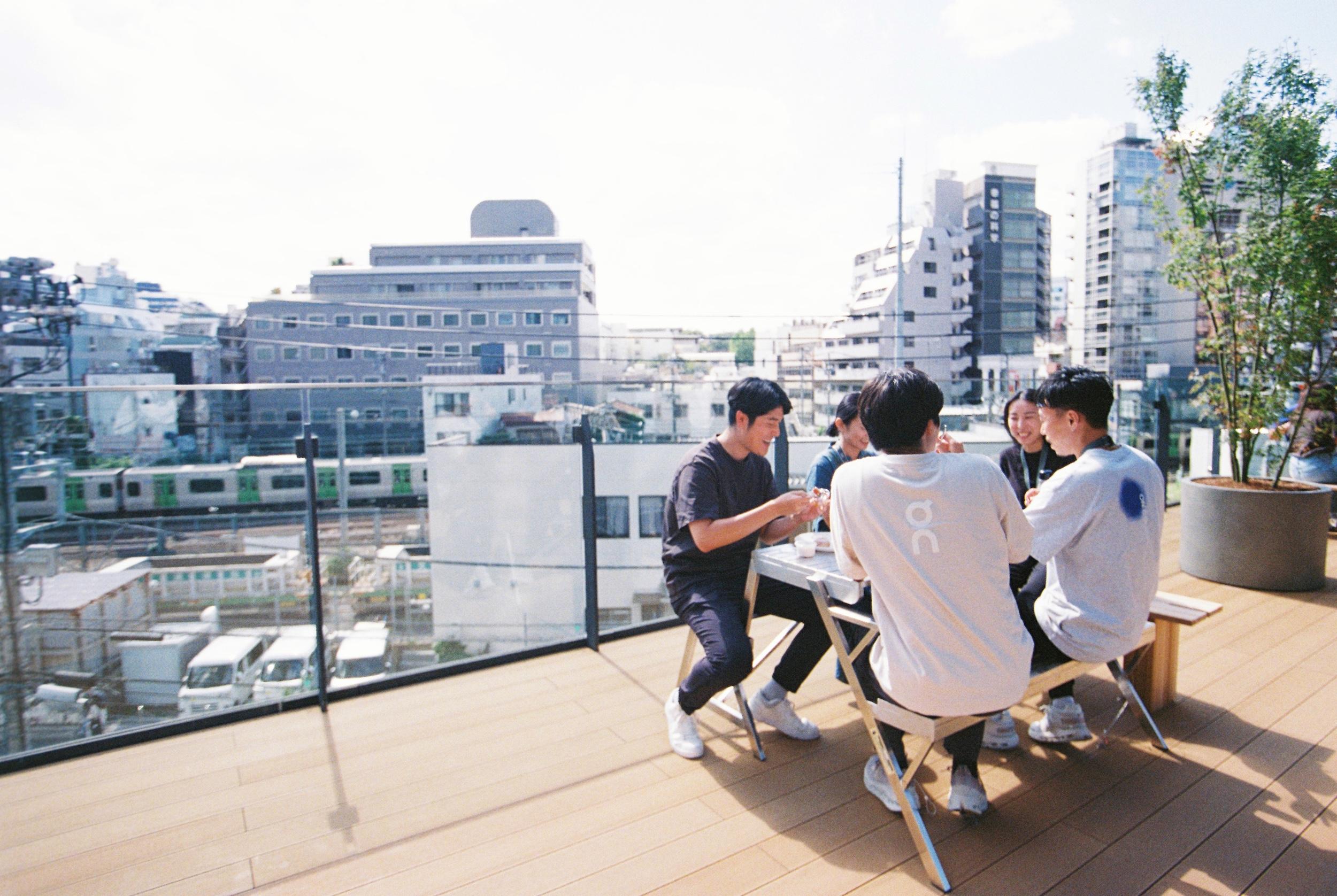 A group of people sit around a small table on a rooftop terrace overlooking city buildings behind a glass railing.