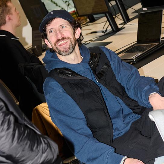 A smiling man wearing a navy baseball cap, blue hoodie and black puffer vest sits at a desk with laptops and monitors behind him.