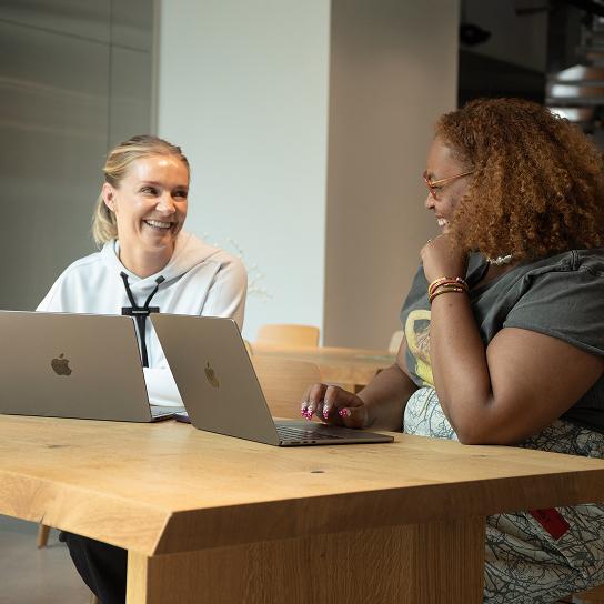 Two people sit at a wooden table with open laptops, smiling and talking to each other.