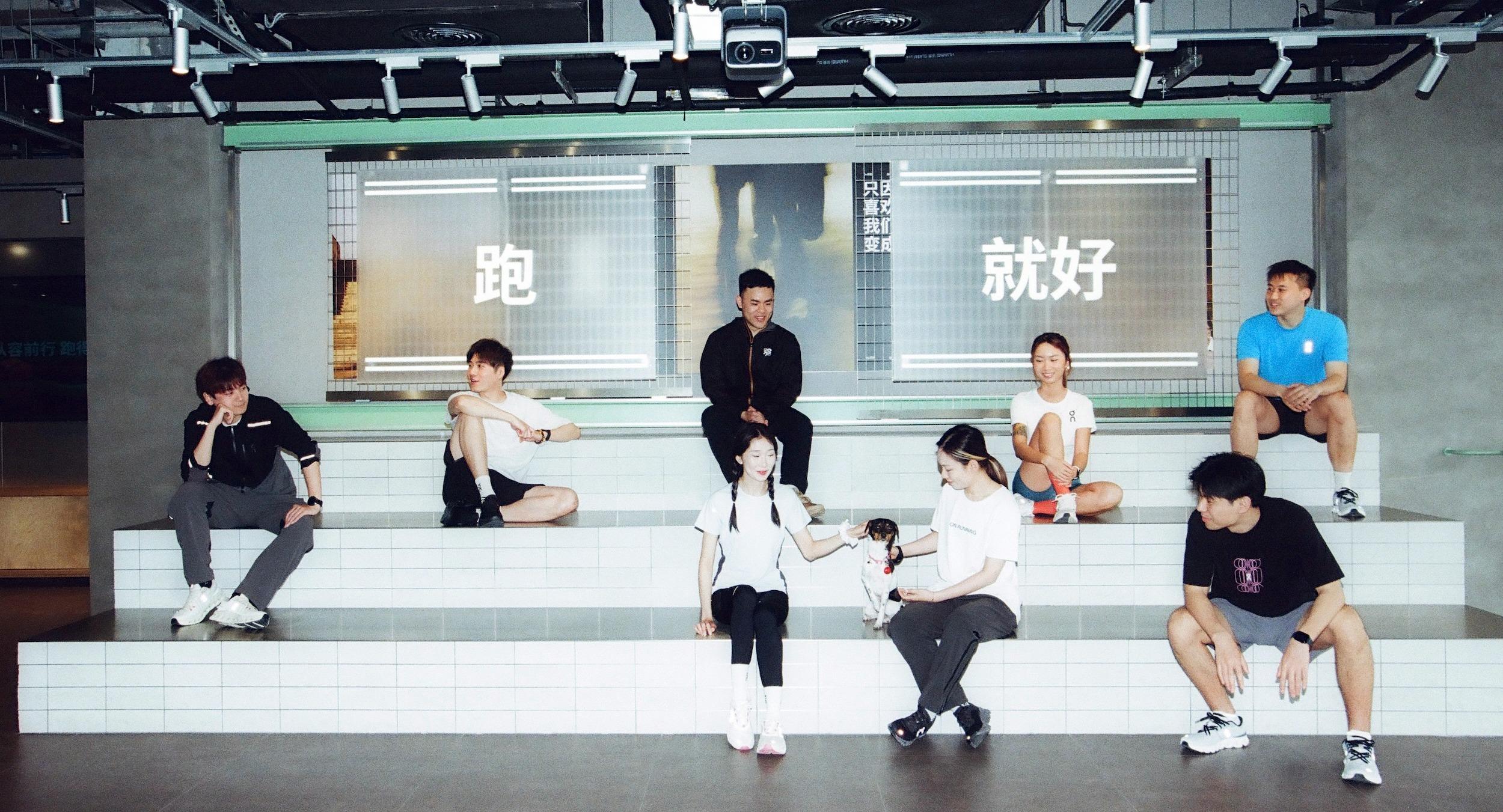 Two young men sit on tiled stepped seating in a modern industrial interior—one in a black hoodie and gray pants and the other in a white T-shirt and black shorts—beneath a lit sign showing the Chinese character "跑".