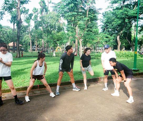 Six people in athletic clothing stand on a paved park path stretching amid trees and grass.