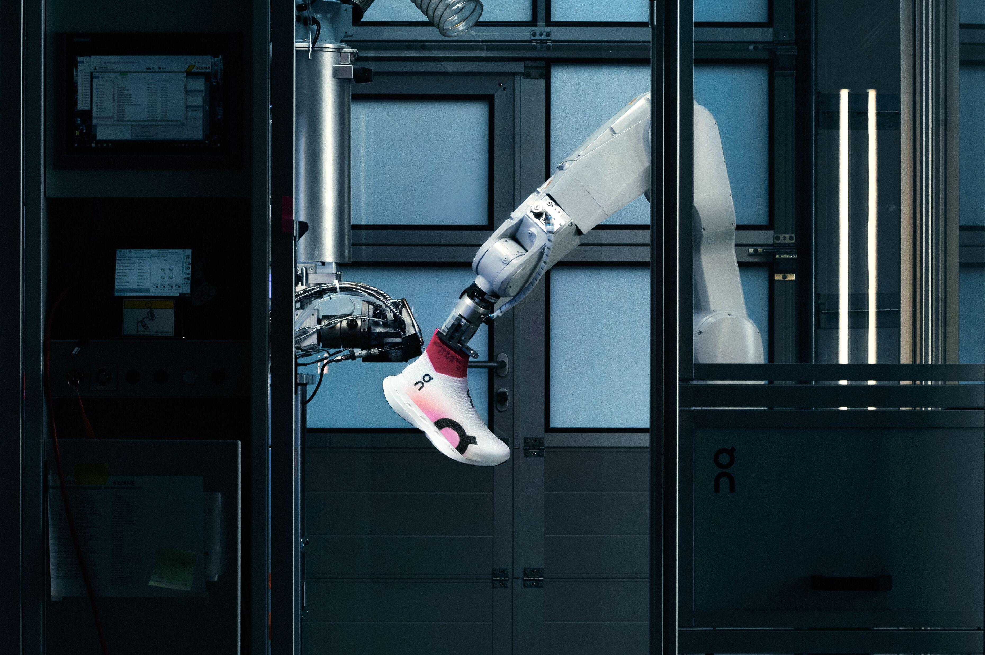 A shoe being held by a motorized robotic arm. It is being sprayed by a second machine with a knit-like webbing to create a lighweight upper material.