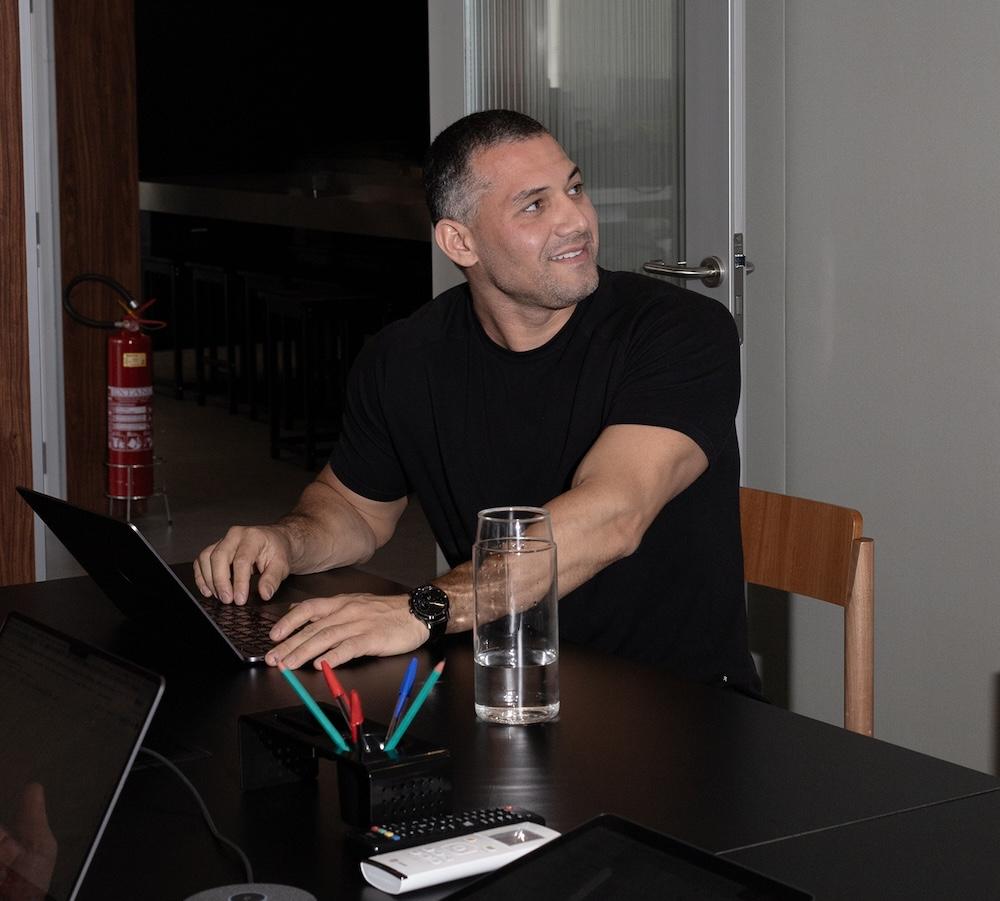 A man in a black T-shirt sits at a table typing on a laptop and looking to the side with a glass of water and pens on the table.