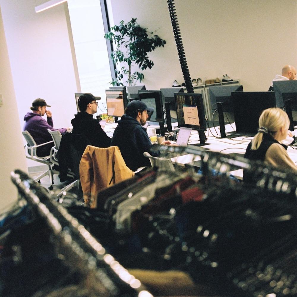 Several people work at desks with desktop computers in an office while a clothing rack with hangers appears in the foreground.