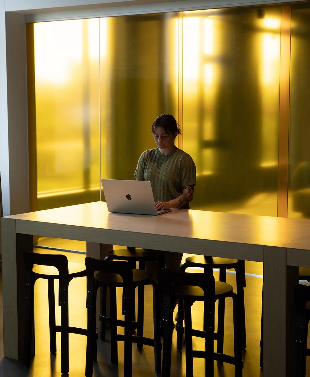 A person with a tattooed forearm using a MacBook at a tall wooden table surrounded by black stools, standing in front of a large glowing yellow wall.