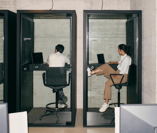 Two people sit separately in adjacent glass-walled work booths using laptops.
