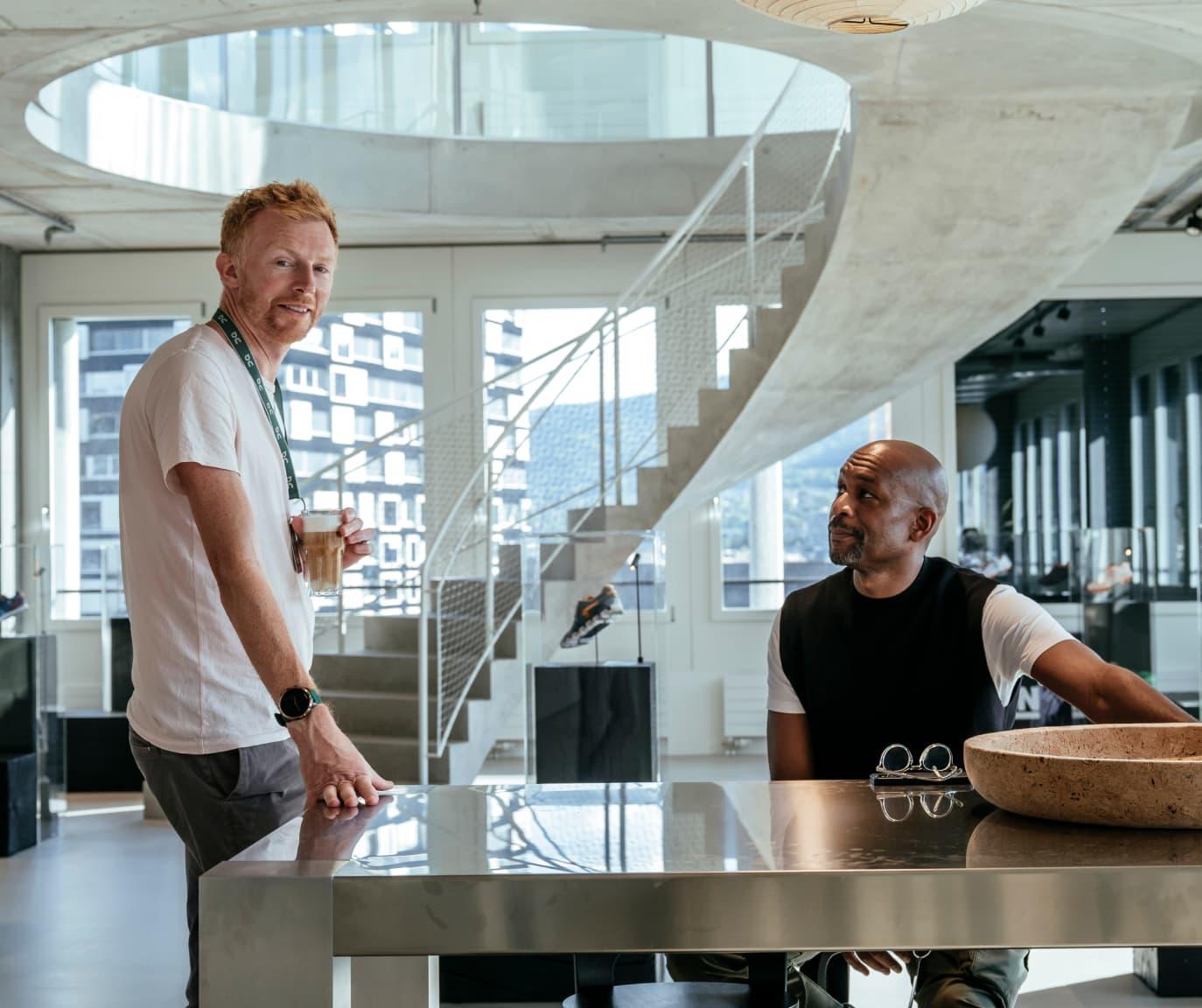 Two men in a modern office lit with natural light and a modern concrete spiral staircase in the background.