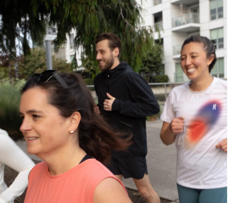 Three adults jogging on a paved path beside apartment buildings—a woman in a coral sleeveless top in the foreground, a man in a black jacket behind her, and a woman in a white shirt smiling to the right.