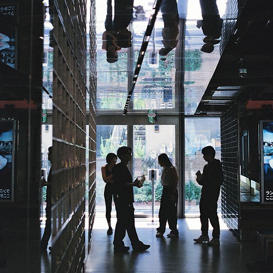 Several silhouetted figures stand in a dim indoor corridor near glass doors, with their reflections visible on the glossy ceiling.