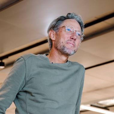 Older man with gray hair and glasses wearing a green sweatshirt, looking slightly upward in a modern indoor space with ceiling lights and panels.