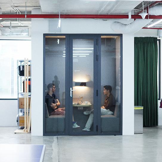 Two people sit facing each other inside a glass-walled office pod in a modern open-plan workspace, with a small table holding a laptop and lamp between them.