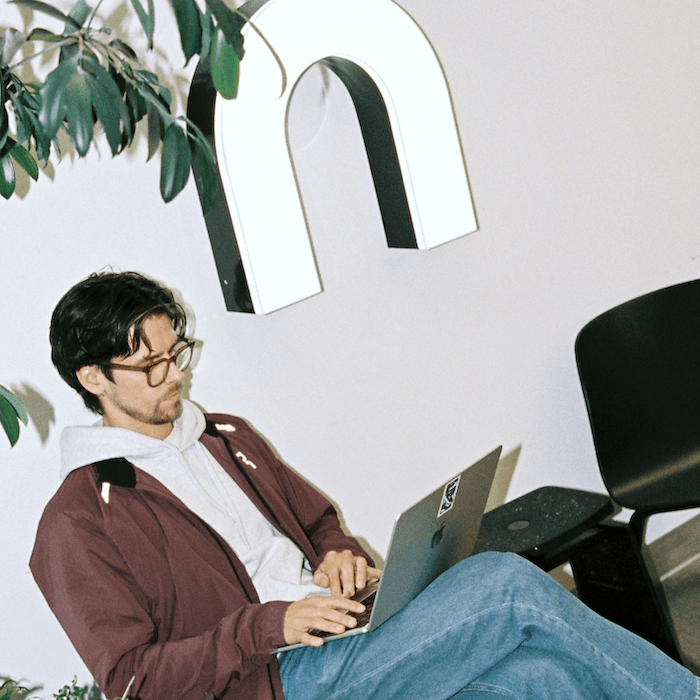 A smiling person wearing glasses and a maroon jacket over a gray hoodie stands leaning on a counter next to a laptop and monitors in an indoor workspace.