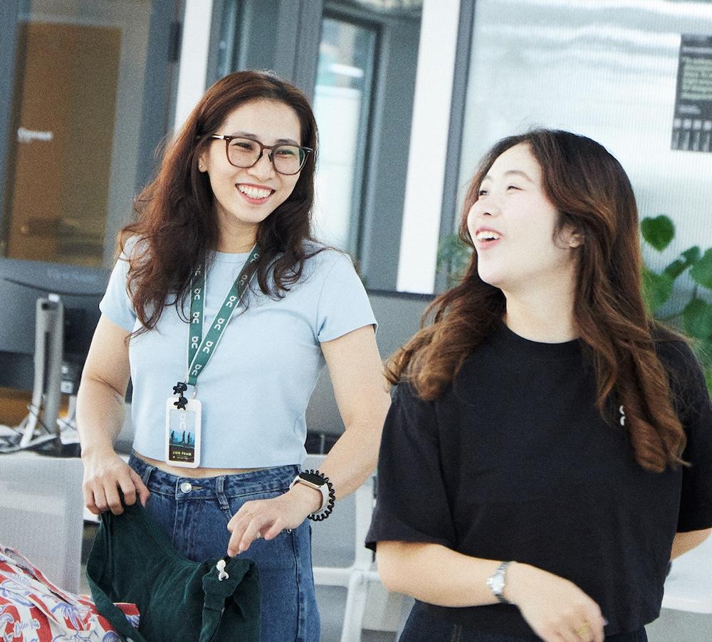 Two women in an office space smile and talk while one, wearing glasses and a lanyard, holds a green bag.