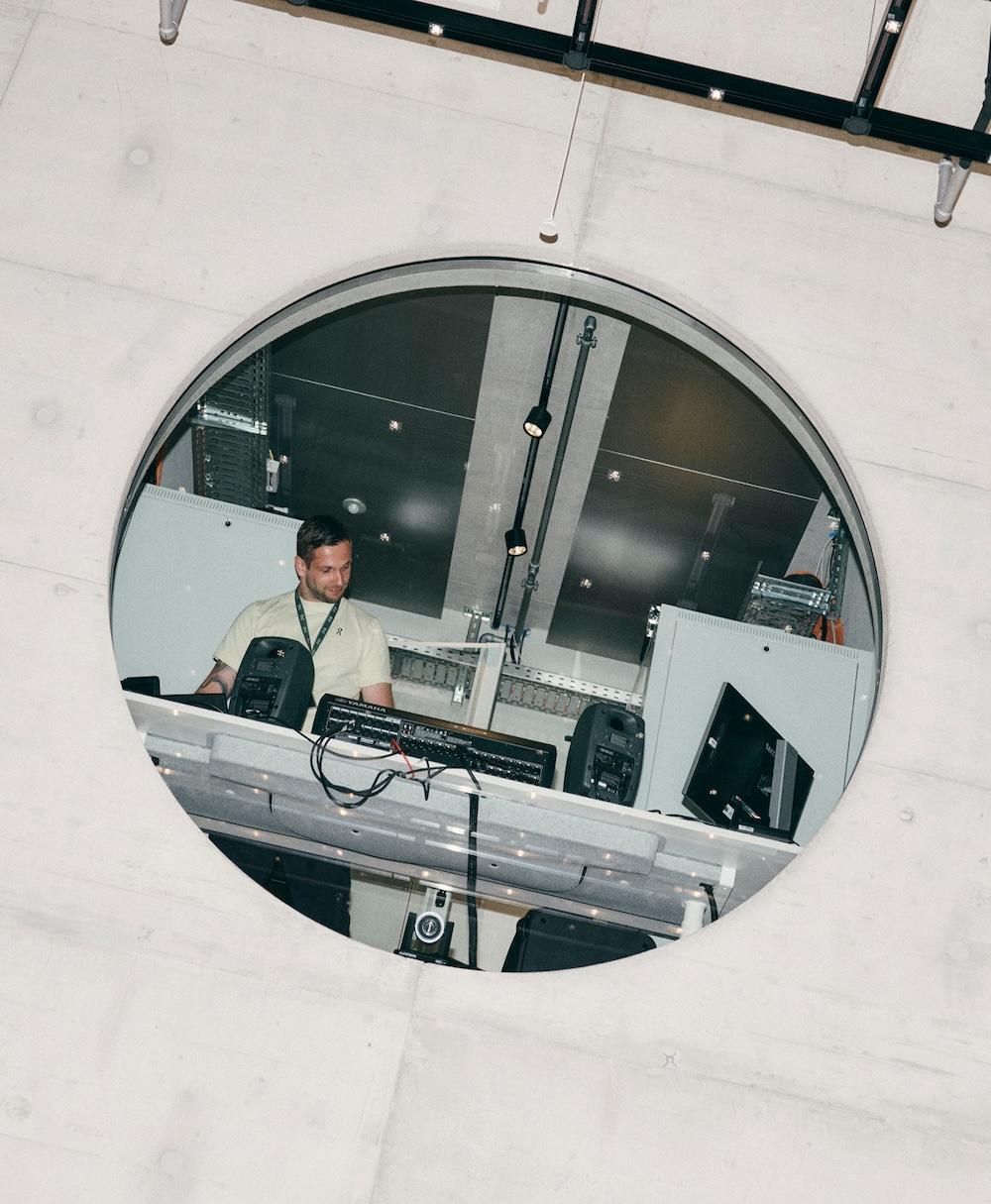 A man stands behind mixers, speakers, and cables in a control room visible through a large circular opening in a concrete wall.