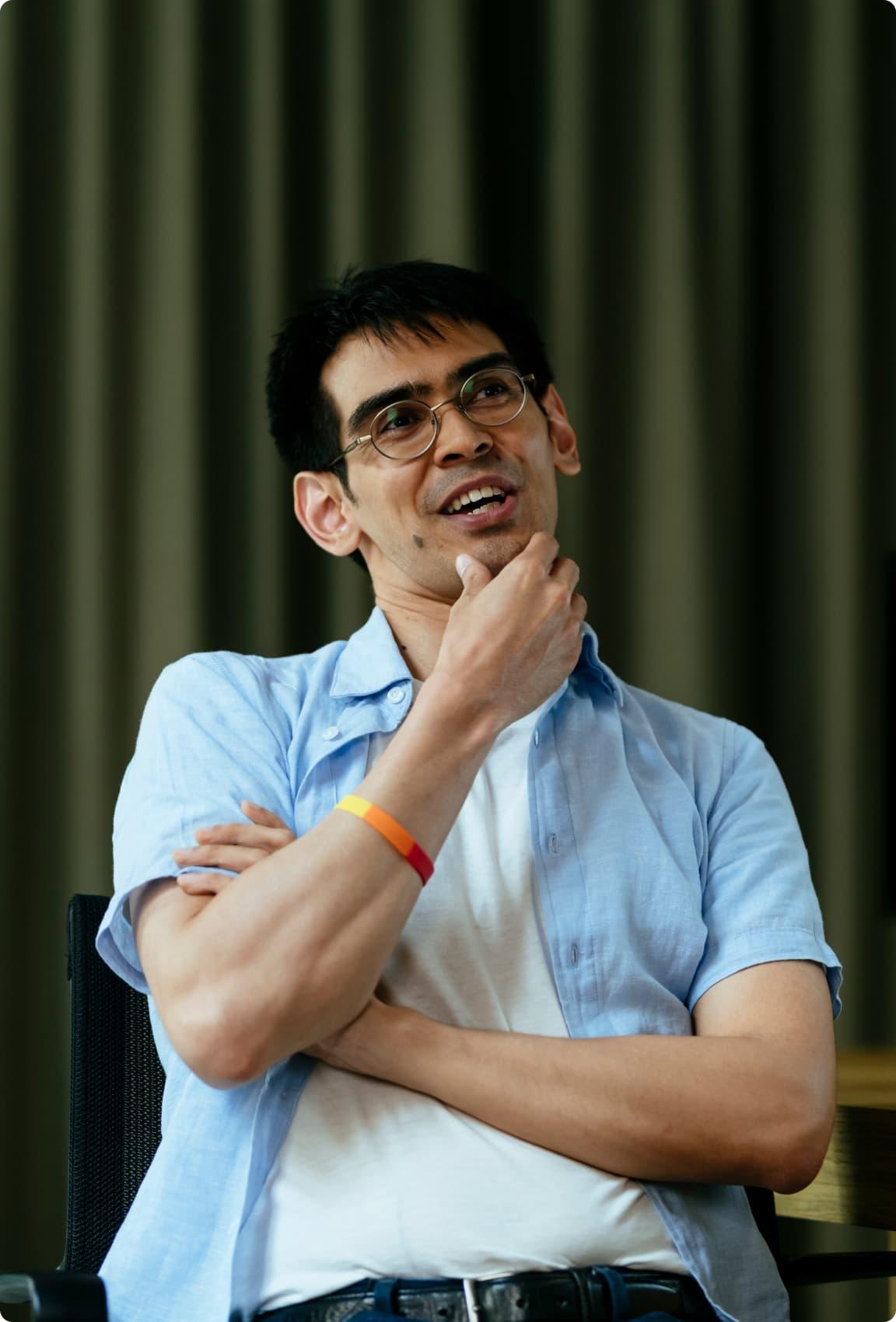 A man in a blue shirt and a white t-shirt with a headset on is standing with his arms crossed and holding his chin looking into the distance