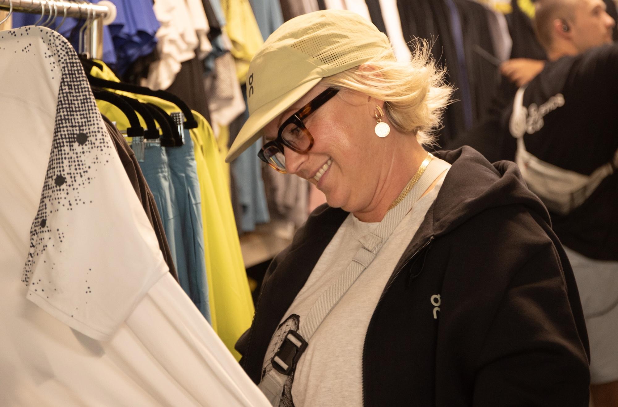 A smiling person wearing a light-colored cap and glasses looks at a white shirt on a clothing rack.