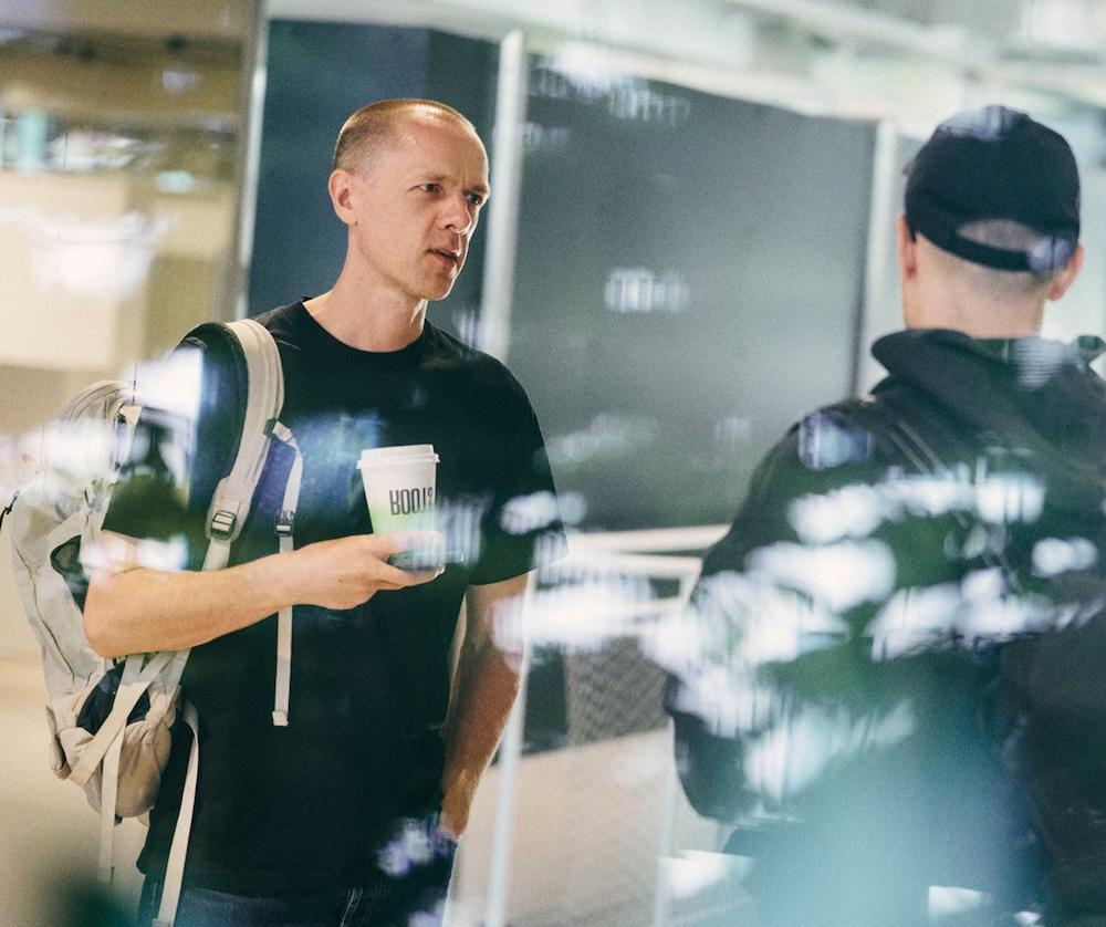 A man wearing a backpack holds a takeaway coffee cup while talking to another person whose back is to the camera.