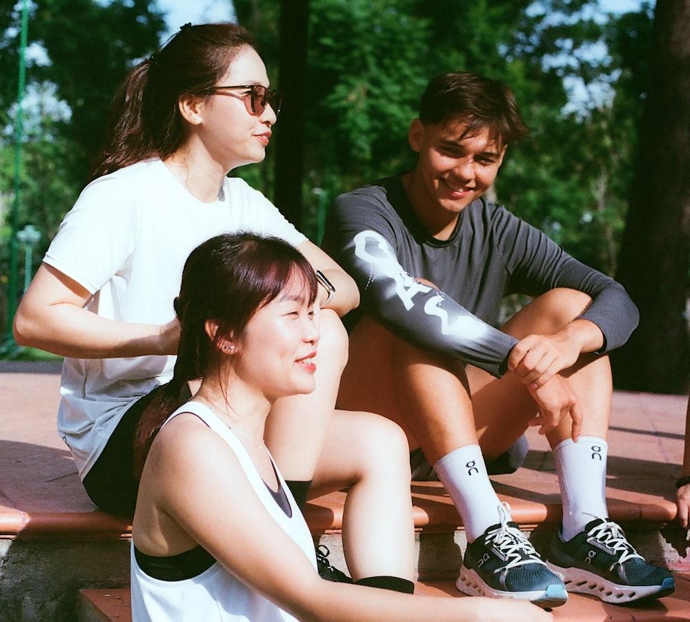 Three people wearing athletic clothing sit on outdoor steps, smiling and talking with trees visible in the background.