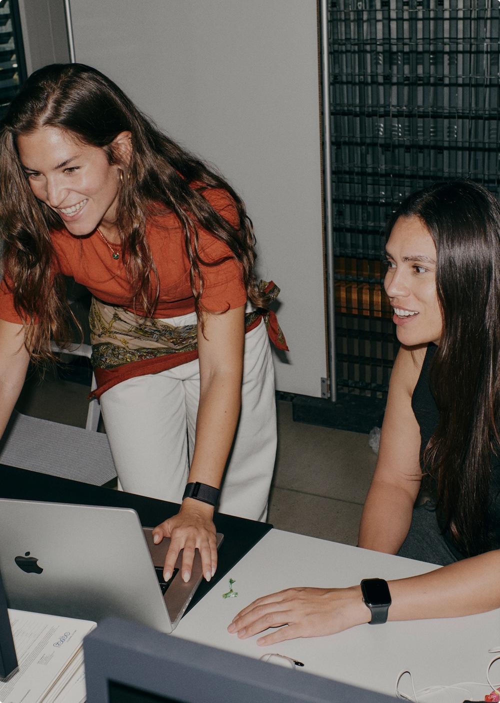 Two women work together at a desk, one leaning over a laptop and smiling while the other sits beside her watching the screen.
