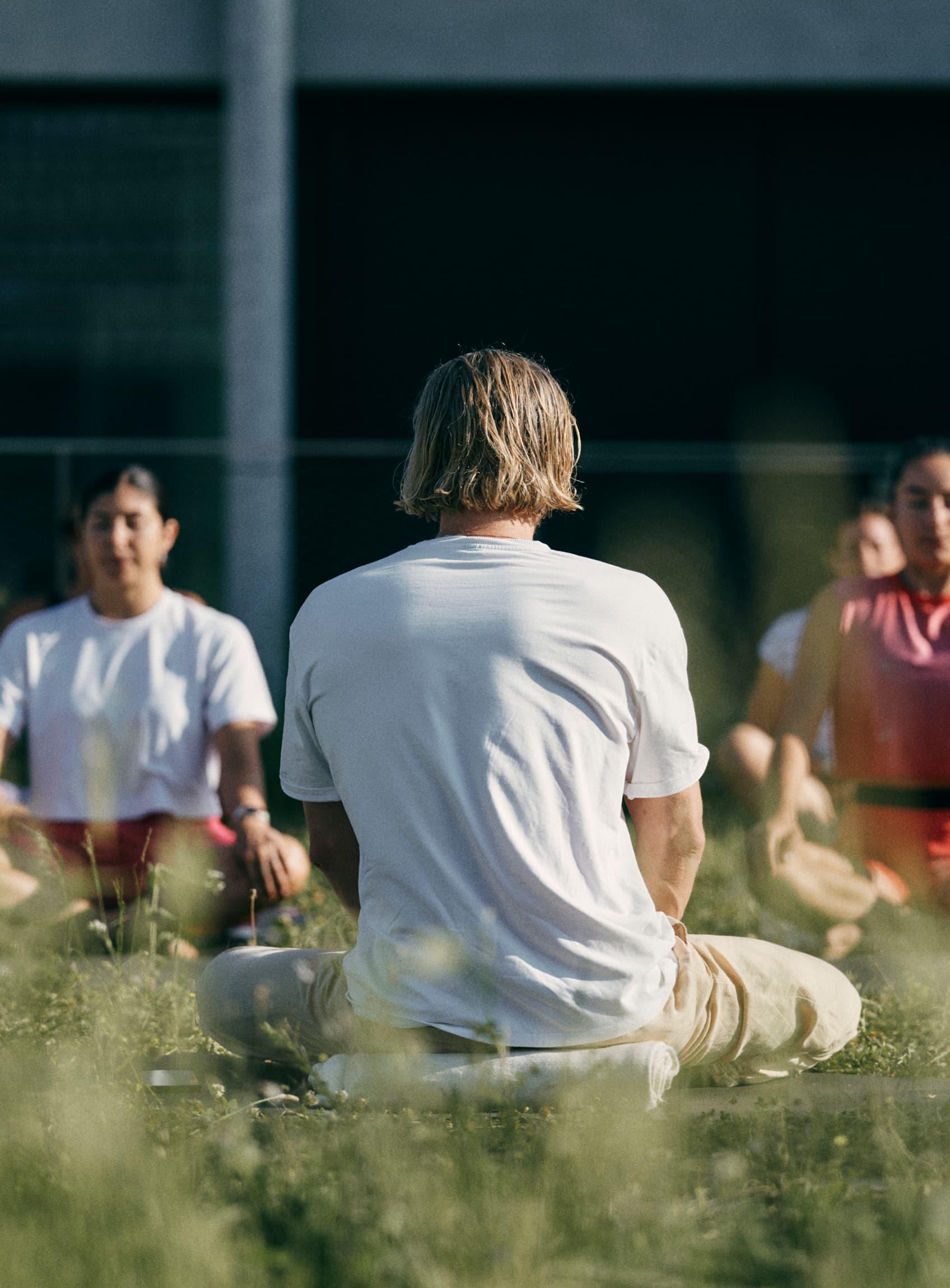 Group meditating cross-legged outdoors in tall grass, facing an instructor in a white shirt.