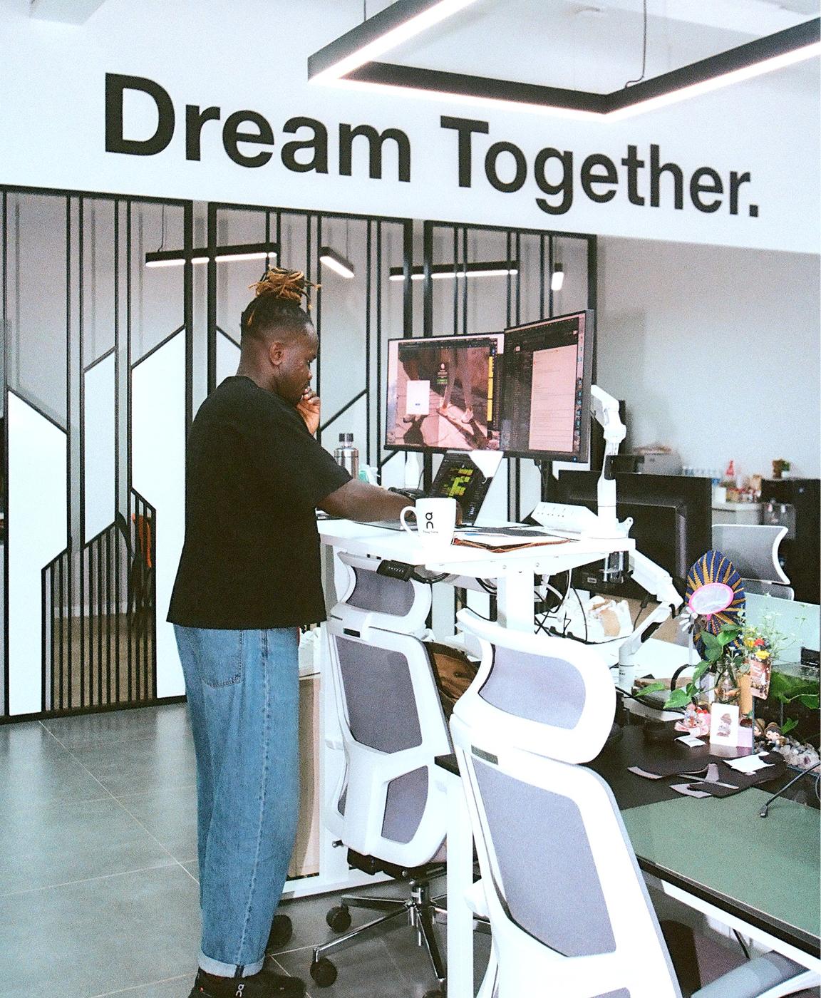 A person stands at a height-adjustable desk using dual monitors in an office with the words "Dream Together." displayed on the wall.