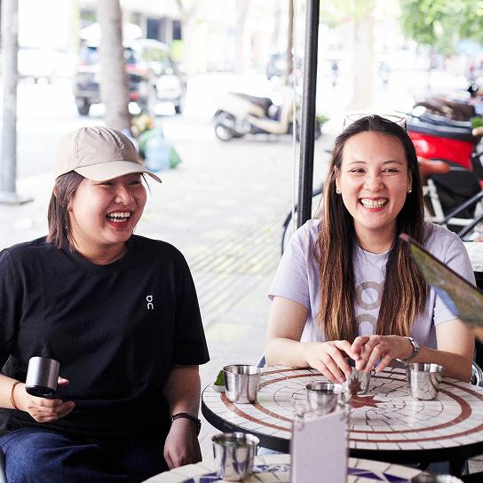Two women sit at a round mosaic outdoor table with metal cups, smiling and laughing—one in a black shirt and beige baseball cap holding a tumbler and the other in a light purple shirt with sunglasses on her head.