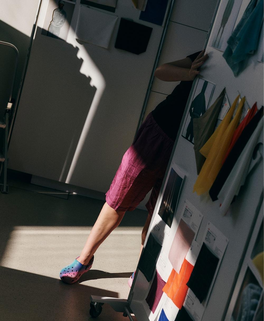 Person in pink shorts and colorful Crocs leans playfully around a fabric sample board in a sunlit design studio.