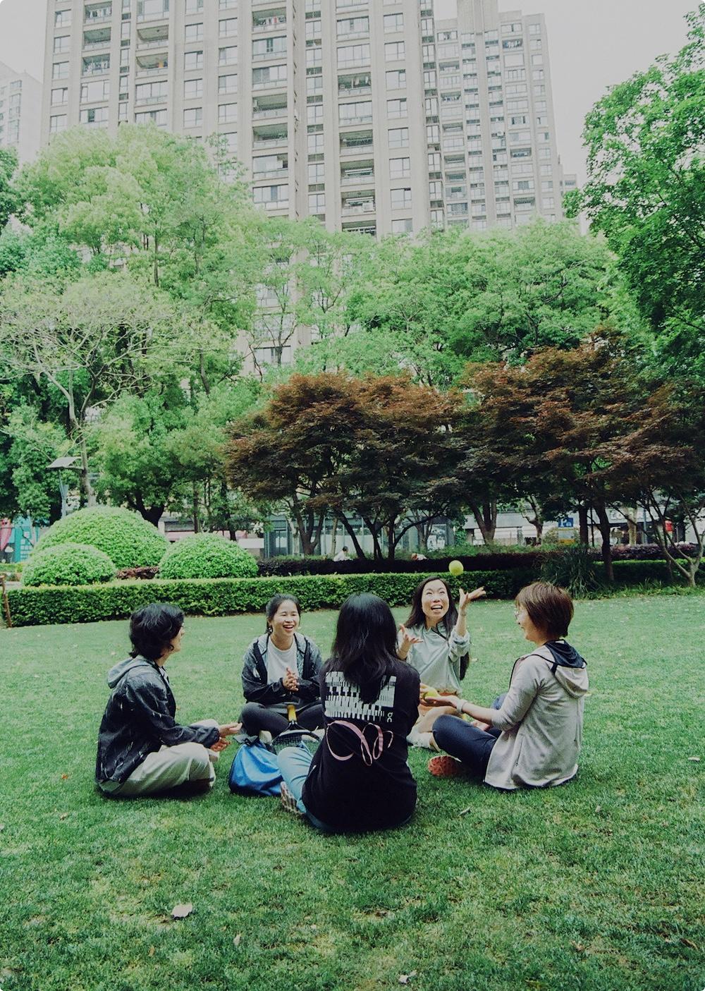Five people sit in a circle on the grass in a city park with trees and a tall apartment building behind them as one person tosses a small ball.