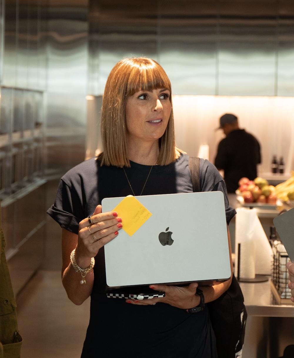 A woman holding a closed silver MacBook with an Apple logo and a yellow sticky note in a stainless-steel kitchen area with fruit and a blurred person in the background.